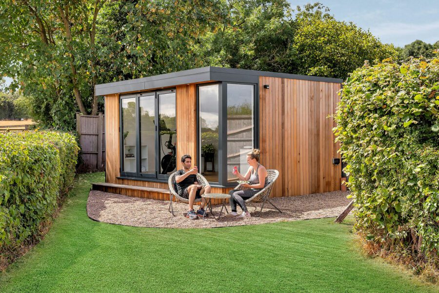 Two people sit outside a modern wooden cabin with large windows, surrounded by lush greenery and hedges, chatting and enjoying beverages after a refreshing workout in their nearby garden gym.