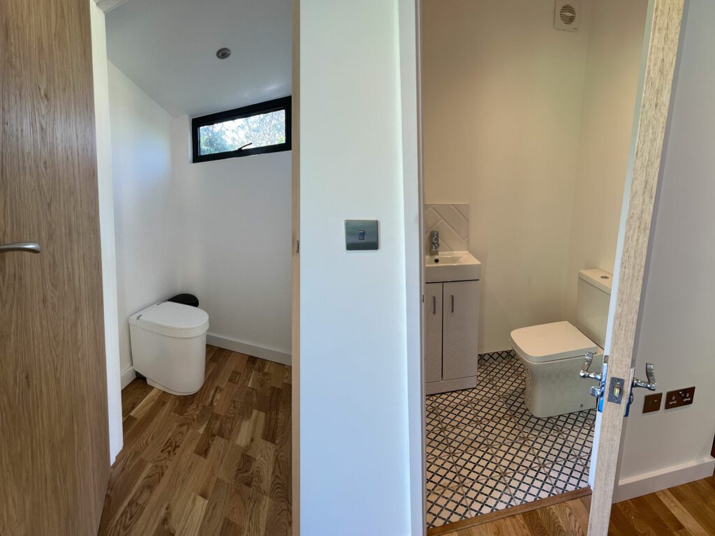 A bathroom with wooden floor and white walls, featuring a white toilet and a small sink with a cabinet under a window. Adjacent is a second room, similar to a garden room with bathroom amenities, complete with another toilet and a small window above.