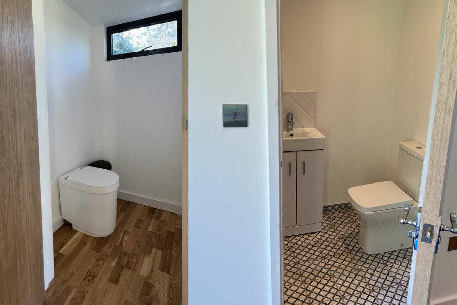 Two separate small rooms: the left contains a toilet with wooden flooring; the right features another toilet, a sink, and patterned tile flooring. Both rooms have white walls and a wooden door frame, complementing a serene garden room with bathroom access.